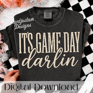 Könnte beinhalten: Ein anthrazitfarbenes Kurzarm-T-Shirt mit einem verwaschenen, cremefarbenen Grafikdruck mit der Aufschrift IT'S GAME DAY darlin. Das Shirt verfügt über einen Rundhalsausschnitt und ein Comfort Colors Markenetikett an der Innenseite des Kragens.