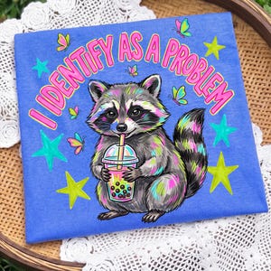 May include: A blue t-shirt featuring a graphic of a raccoon holding a bubble tea cup. The text "I IDENTIFY AS A PROBLEM" is printed in pink above the raccoon. The design includes colorful butterflies and bright green stars surrounding the raccoon.