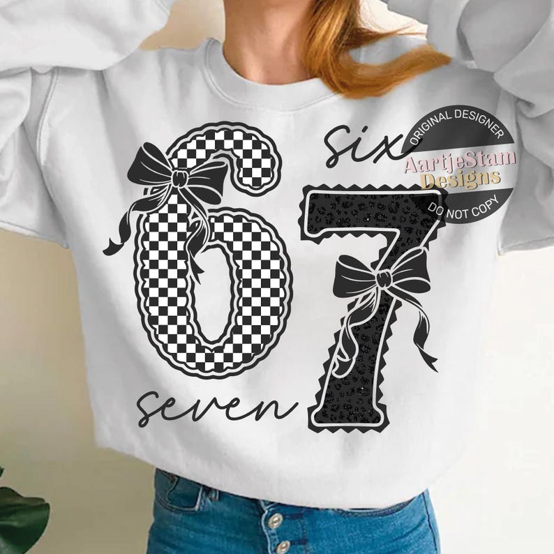 May include: White sweatshirt with the numbers "67" in black and white checkered and leopard print patterns. The word "six" is above the number 6, and the word "seven" is below the number 67. Each number has a black bow.