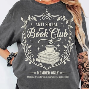 Puede incluir: Camiseta gris oscuro con un gráfico crema. El gráfico presenta el texto "ANTI SOCIAL Book Club" dentro de un marco decorativo con hojas y estrellas. Debajo, una pila de libros con una taza de café. El texto "MEMBER ONLY" está debajo.