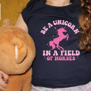 May include: A navy blue t-shirt with a pink unicorn graphic and the text "Be a unicorn in a field of horses".