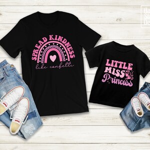 May include: Two black t-shirts with pink text and graphics. The larger shirt says "Spread Kindness like confetti" with a pink rainbow and hearts. The smaller shirt says "Little Miss Princess" with a pink crown and a princess silhouette.