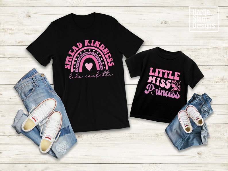 May include: Two black t-shirts with pink text and graphics. The larger shirt says "Spread Kindness like confetti" with a pink rainbow and hearts. The smaller shirt says "Little Miss Princess" with a pink crown and a princess silhouette.
