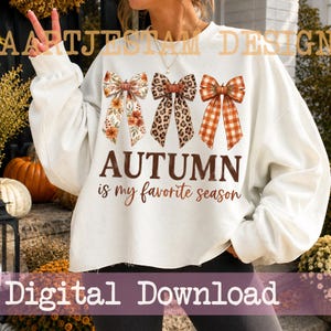 Könnte beinhalten: Ein weißes Langarm-Sweatshirt mit drei dekorativen Schleifen in Blumen-, Leoparden- und orangefarbenen Karomustern. Der Text "AUTUMN is my favorite season" ist in Braun unter den Schleifen aufgedruckt. Das Kleidungsstück hat einen Rundhalsausschnitt.