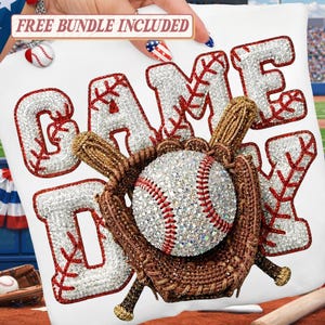 以下が含まれることがあります： 野球をテーマにしたデザインで「GAME DAY」の文字がプリントされた白いTシャツ。文字は赤いステッチで縁取られ、ラインストーンが詰められています。野球ボールとミットが中央にあります。
