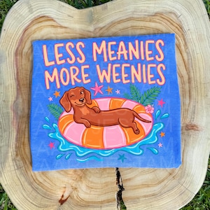 Könnte beinhalten: Blaues T-Shirt mit dem Text "LESS MEANIES MORE WEENIES" in Orange und Pink. Ein Cartoon-Dackel schwimmt auf einem rosa-orangenen Schwimmring. Das Design umfasst Sterne, Wasserspritzer und tropische Blätter.