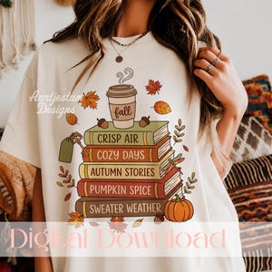 Könnte beinhalten: Ein cremefarbenes T-Shirt mit einer Grafik von fünf gestapelten Büchern mit den Aufschriften "CRISP AIR", "COZY DAYS", "AUTUMN STORIES", "PUMPKIN SPICE" und "SWEATER WEATHER". Ein mit "fall" beschrifteter Kaffeebecher steht obenauf, umgeben von Blättern, Eicheln und einem Kürbis.