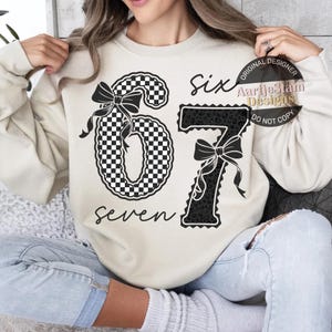 May include: A cream-colored sweatshirt featuring the numbers "67" in a black and white checkered pattern and a black leopard print. The word "six" and "seven" are written in black script. The design includes black bows.