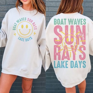 Op de afbeelding: Crèmekleurige sweatshirt met de zinnen "Boat Waves Sun Rays Lake Days" en een gele smiley-afbeelding. De achterkant van de sweatshirt herhaalt de zinnen en smiley in een verticale lay-out.
