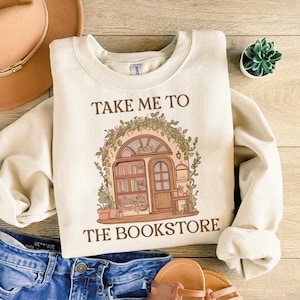 Könnte beinhalten: Ein cremefarbener Pullover mit dem Text "Take me to the bookstore" auf der Vorderseite. Der Text ist in braunen Buchstaben. Die Grafik zeigt eine Comic-Illustration eines Buchladens mit einer Tür, Bücherregalen und Pflanzen.
