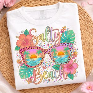 Könnte beinhalten: Ein weißes T-Shirt mit einer bunten Grafik aus einer Sonnenbrille mit Leopardenmuster. Die Gläser zeigen einen Strandsonnenuntergang mit Palmen und Wellen. Der Schriftzug Salty Beach ist in Schreibschrift gehalten, umgeben von Hibiskusblüten.