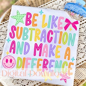 Könnte beinhalten: Ein weißes T-Shirt mit dem bunten Schriftzug BE LIKE SUBTRACTION AND MAKE A DIFFERENCE. Das Design enthält einen grünen, gepunkteten Stern, eine leuchtend rosa Schleife, ein rosa, gepunktetes Smiley-Gesicht und kleine gelbe und blaue Sterne.