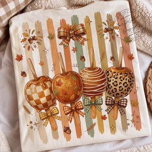 May include: A cream-colored t-shirt featuring a graphic of four caramel apples on sticks against a background of vertical brushstrokes in muted autumn colors. The apples have checkered, leaf, striped, and leopard patterns with plaid bows.