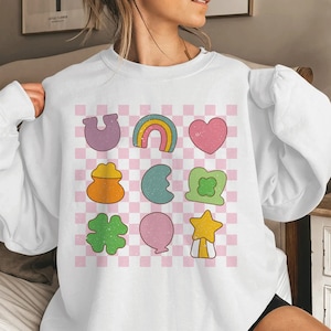 Könnte beinhalten: Weißes Sweatshirt mit einem Raster aus farbenfrohen Grafiken im Retro-Stil. Die Grafiken umfassen ein Hufeisen, einen Regenbogen, ein Herz, einen Goldtopf, einen Halbmond, ein vierblättriges Kleeblatt, einen Ballon und einen Stern. Der Hintergrund ist ein rosa-weißes Karomuster.