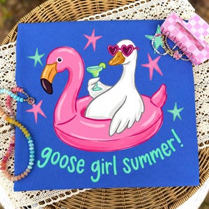 Könnte beinhalten: Blaues T-Shirt mit einem Flamingo und einer Gans in einem rosa Schwimmring, beide mit Sonnenbrillen und Cocktails. Der Text "goose girl summer!" ist in grüner Schrift geschrieben. Rosa und grüne Sterne umgeben das Bild.