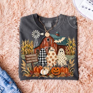 Könnte beinhalten: Ein dunkelgraues Sweatshirt mit Rundhalsausschnitt und einem Aufdruck einer roten Scheune, einer Windmühle, Maiskolben und drei Geistern. Die Geister haben Karomuster, Blumenmuster und Punkte. Ein Holzschild trägt die Aufschrift PUMPKINS HAY-RIDES CORN MAZE.