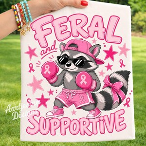 Könnte beinhalten: Eine weiße Stoffoberfläche mit einem Cartoon-Waschbären, der eine rosa Baseballkappe, eine schwarze Sonnenbrille, rosa Boxhandschuhe und rosa Shorts trägt. Der Waschbär hat eine rosa Schleife am Schwanz. Text FERAL und SUPPORTIVE mit rosa Sternen.