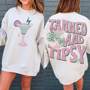 Peut inclure: Un sweat-shirt blanc avec un graphique de margarita verte sur le devant et le texte "Tanned and Tipsy" en rose et vert au dos.