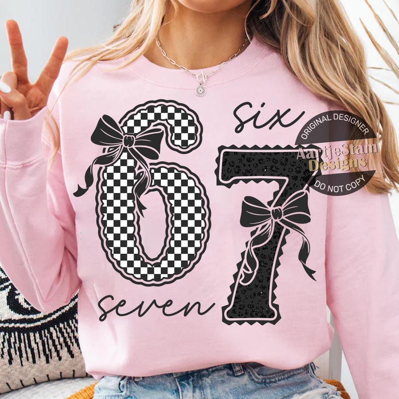 May include: A light pink sweatshirt with the numbers "67" in a black and white checkered pattern and black leopard print. The word "six" is written above the number 6 and the word "seven" is written below the number 7.