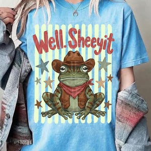 May include: A light blue t-shirt with the text "Well. Sheeeyit" in red letters. The shirt features a frog wearing a cowboy hat, vest, and red bandana. The frog is surrounded by stars and vertical stripes.