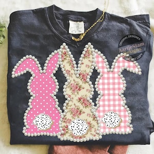 Könnte beinhalten: Dunkelblaues T-Shirt mit drei Osterhasen-Designs. Die Hasen sind mit rosa Punkten, floralen Mustern und rosa Vichy-Karo verziert. Jeder Hase ist mit Kunstperlen umrandet und hat einen Schwanz mit Dalmatiner-Print. Das Shirt hat den Text "AartjeStam Designs".