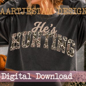 Könnte beinhalten: Ein anthrazitfarbenes Sweatshirt mit Rundhalsausschnitt und dem Text He's HUNTING in College-Schriftart. Das Wort HUNTING ist mit einem grünen, braunen und beigen Tarnmuster gefüllt und weist sichtbare Ziernähte an den Buchstabenrändern auf.