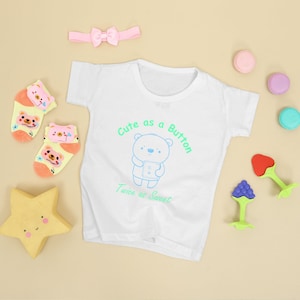 Op de afbeelding: Een wit babypakje met een schattige cartoonbeer en de tekst "Cute as a Button Twice as Sweet". Het pakje is omringd door babysokken, een hoofdband en bijtringen.