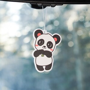 May include: A white car air freshener with a cartoon panda holding a piece of bamboo. The panda has black eyes, a black nose, and black ears. The panda is wearing a black and white striped shirt.