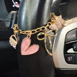 Op de afbeelding: Een goudkleurige bedelarmband met een roze hartvormige hanger, een gouden strik, een roze vlinderbedel en een kleine handtasbedel. De armband hangt over een zwart stuurwiel met zichtbare knoppen met de tekst RESUME en SET.