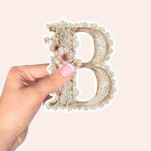 May include: A decorative sticker of the letter "B", featuring a vintage design. The letter is embellished with lace and soft pink flowers. A hand with a pink manicure holds the sticker.