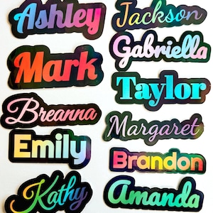 May include: A collection of ten individual name stickers featuring holographic, multi-coloured lettering on a black background. The names displayed are Ashley, Jackson, Mark, Gabriella, Breanna, Taylor, Margaret, Emily, Brandon, and Kathy, and Amanda.