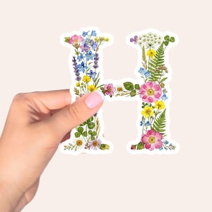 May include: A sticker in the shape of the letter "H" is filled with a variety of colorful wildflowers. The sticker is held by a hand with pink nail polish against a light background. The flowers include pink, yellow, blue, and white blooms.