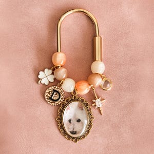 May include: A gold-toned U-shaped bag charm featuring peach and cream-coloured beads, a white four-leaf clover charm, a circular pendant with the letter D, a star-shaped charm, and an oval frame containing a photograph of a white dog.