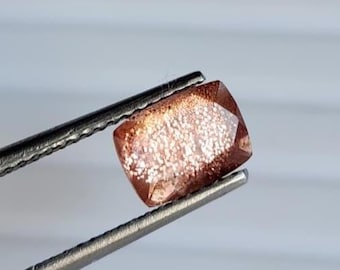 May include: A faceted, rectangular sunstone gemstone held by tweezers. The gem has a warm, reddish-brown hue with sparkling inclusions, creating a shimmering effect. The stone is cut with rounded corners and is ready for jewelry making.