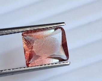 May include: A square-cut gemstone held by tweezers. The gem has a peach and clear color with a glittery appearance. The edges of the gem are a darker shade of red. The background is white.