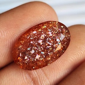 May include: Oval-shaped gemstone with a warm, reddish-brown hue. The stone is speckled with iridescent inclusions that shimmer with colors like pink, blue, and gold. The gem is held in a hand, showcasing its polished surface and unique internal patterns.