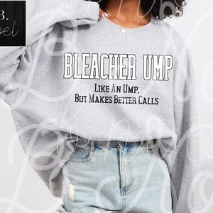 Op de afbeelding: Grijs sweatshirt met de tekst "Bleacher Ump, Like an Ump, But Makes Better Calls" in zwart gedrukt.