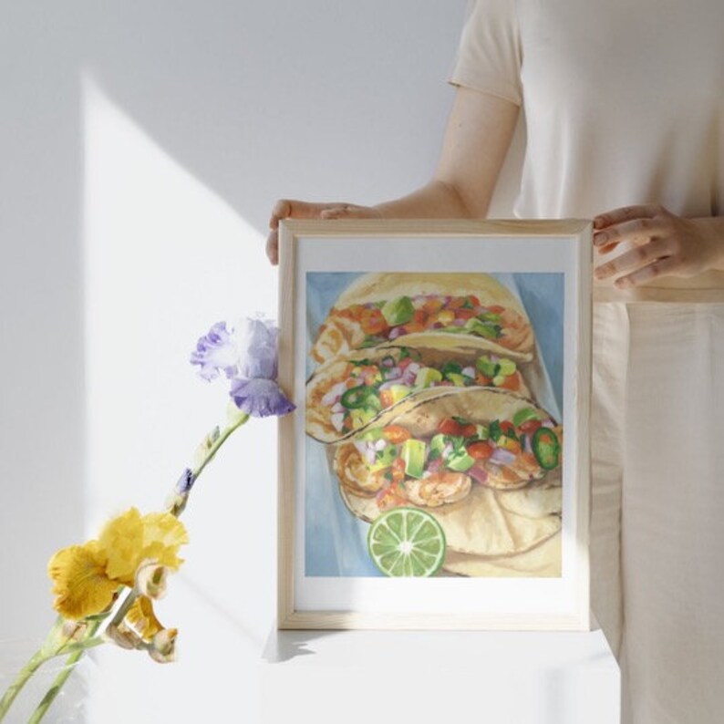Może przedstawiać: Namalowany obraz w ramie przedstawiający trzy taco z krewetkami, awokado i pomidorem. Taco leżą na białym talerzu z plasterkiem limonki.