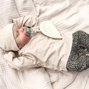 Pode incluir: Um recém-nascido usando um chapéu bege, camisa de manga comprida e calças com estampa de leopardo, deitado em um cobertor bege. O bebê tem uma chupeta e um item decorativo em forma de asa. A paleta de cores é suave e neutra.
