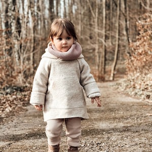 Könnte beinhalten: Ein kleines Kind trägt einen beigefarbenen Pullover mit einem rosa Schalkragen-Schal und braunen Stiefeln und geht auf einem Weg in einem Wald.