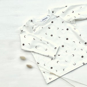 Könnte beinhalten: Ein weißes Langarmshirt mit einem Halbmondmuster. Das Shirt hat einen Rundhalsausschnitt und ist aus einem weichen, bequemen Stoff gefertigt.