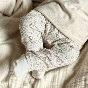 Könnte beinhalten: Nahaufnahme der Beine und Füße eines Babys. Das Baby trägt eine Hose mit beige-weißem Leopardenmuster und ein hellbeige Langarm-Oberteil. Weiße Socken bedecken die Füße des Babys. Das Baby liegt auf einer beigen Decke.