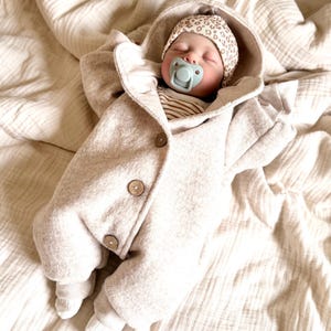 Könnte beinhalten: Ein schlafendes Baby in einem beigen Kapuzenmantel mit Holzknöpfen. Das Baby trägt auch einen gemusterten Hut und hat einen Schnuller im Mund. Der Mantel ist aus einem weichen Material gefertigt und liegt auf einer beigen Decke.