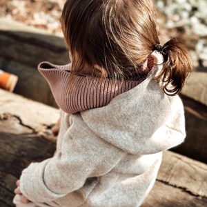 May include: A young child wearing a light beige hooded sweater with a pink knit scarf. The child is sitting on a wooden log.