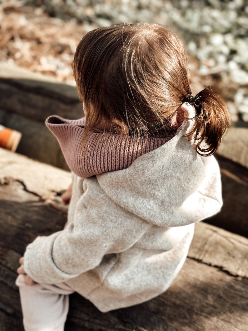 May include: A young child wearing a light beige hooded sweater with a pink knit scarf. The child is sitting on a wooden log.