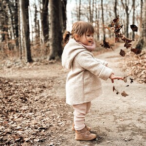 May include: A young child wearing a beige coat and pink leggings throws leaves in the air while standing on a path in a forest.