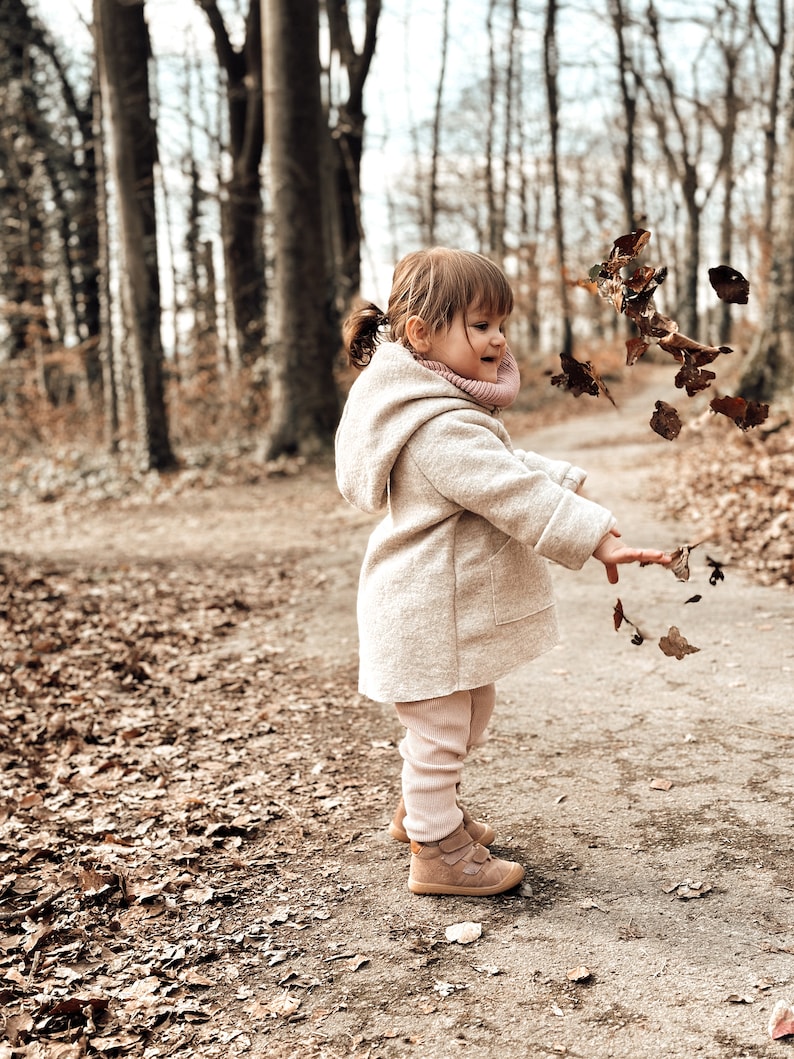 May include: A young child wearing a beige coat and pink leggings throws leaves in the air while standing on a path in a forest.