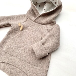 May include: A brown hooded sweatshirt with a white lining featuring a pattern of small moons and dots. The sweatshirt has a kangaroo pocket and a curved hem.
