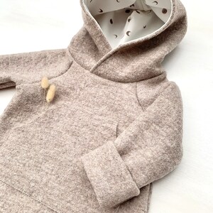 May include: A beige hooded sweatshirt with a white lining featuring a pattern of small brown moons. The sweatshirt has a kangaroo pocket and long sleeves with cuffed ends.