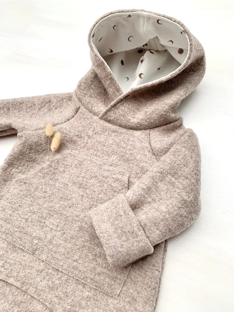 May include: A beige hooded sweatshirt with a white lining featuring a pattern of small brown moons. The sweatshirt has a kangaroo pocket and long sleeves with cuffed ends.